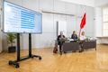 Weitwinkelaufnahme der Pressekonferenz mit den Vortragenden rechts und einem Bildschirm, der eine Präsentationsfolie zeigt, links im Bild.